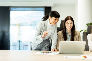 Çağdaş bir ofis ortamında, iki kadın keyifli bir sohbete girişiyor, bir dizüstü bilgisayar üzerinde çalışırken fikir alışverişinde bulunuyor ve gülümsüyor, pozitif ve işbirlikçi bir atmosfer yaratıyor..