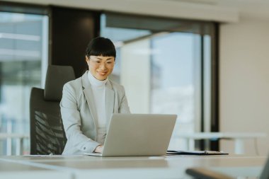 Açık gri takım elbiseli çalışkan bir Japon kadının laptopuna odaklandığı parlak ve geniş çalışma alanı, odayı doğal ışık kapladıkça azim ve profesyonellik gösteriyor..