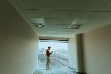 Modern bir binanın üst katında, gözlüklü bir adam tek başına ayakta duruyor, aşağıdaki hareketli şehrin panoramik manzarasının keyfini çıkarırken mobil ekranına odaklanıyor..