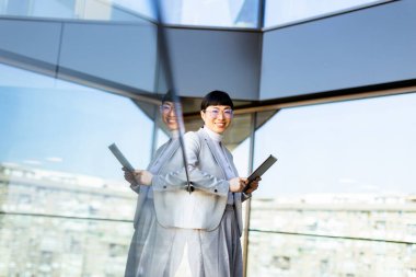 Kendine güvenen bir girişimci elinde tabletle parlak, çağdaş bir ofiste duruyor. Büyüleyici şehir silüeti büyük cam pencerelerden görülebilir..