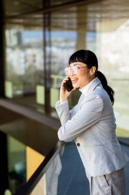 Modern ofis ortamında, şık takım elbiseli kendine güvenen Asyalı bir kadın telefonda konuşurken geniş bir gülümseme sergiliyor, yansıması şehrin cam zemininde görülebiliyor..