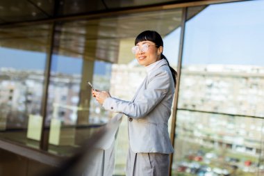 Parlak güneş ışığı şık takım elbiseli kendine güvenen Asyalı bir kadını aydınlatıyor, akıllı telefonuyla hareketli bir şehir ortamında çağdaş mimarinin arka planına karşı tebessüm ederken gülümsüyor..