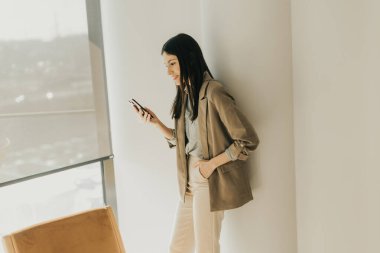 Akıllı telefonuna odaklanmış bir kadın duvara yaslanırken, parlak güneş ışığı geniş pencerelerden içeri akıyor. Havalı kıyafetler giyerken sükunet saçıyor..