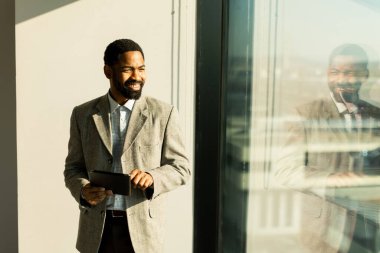 İyi giyimli bir adam parlak bir ofis alanında tablet kullanırken gülümser. Güneş ışığı sıcak bir parıltı saçar ve üretken bir anın tadını çıkarırken odaklanmış yüz ifadesini vurgular..