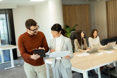 Işıl ışıl bir ofiste iki profesyonel iş birliği içinde hareketli bir değişim gerçekleşiyor. Arka planda, iş arkadaşları dizüstü bilgisayarlarına odaklanarak takım çalışması ve yenilik atmosferini artırıyorlar..