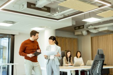Işıl ışıl bir ofiste iki profesyonel iş birliği içinde hareketli bir değişim gerçekleşiyor. Arka planda, iş arkadaşları dizüstü bilgisayarlarına odaklanarak takım çalışması ve yenilik atmosferini artırıyorlar..