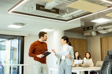 Işıl ışıl bir ofiste iki profesyonel iş birliği içinde hareketli bir değişim gerçekleşiyor. Arka planda, iş arkadaşları dizüstü bilgisayarlarına odaklanarak takım çalışması ve yenilik atmosferini artırıyorlar..