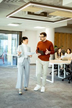 Işıl ışıl bir ofiste iki profesyonel iş birliği içinde hareketli bir değişim gerçekleşiyor. Arka planda, iş arkadaşları dizüstü bilgisayarlarına odaklanarak takım çalışması ve yenilik atmosferini artırıyorlar..