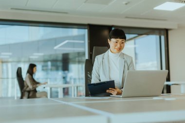 Asyalı iş kadını, geniş pencereli şık bir ofisteki görevlere dikkatle odaklanarak dinamik ve dinamik bir atmosfer yaratıyor.