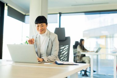 Şık bir ofiste, özel dikim takım elbiseli odaklı Asyalı bir kadın dizüstü bilgisayarında, bir meslektaşı yakınlarda çalışırken gözlüğünü ayarlıyor, modern bir ortamda verimlilik sergiliyor..