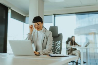 Şık bir ofiste, özel dikim takım elbiseli odaklı Asyalı bir kadın dizüstü bilgisayarında, bir meslektaşı yakınlarda çalışırken gözlüğünü ayarlıyor, modern bir ortamda verimlilik sergiliyor..