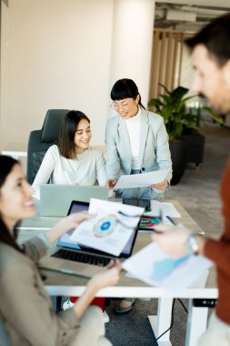 Çağdaş bir çalışma alanında, bir ekip canlı bir tartışma içine girer, büyük bir masa etrafında fikirler ve belgeler paylaşır. İfadeleri coşku ve takım çalışmasını yansıtıyor.