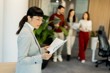 Gösterişli bir ofiste önemli belgeleri inceleyen odaklanmış bir profesyonel, meslektaşları arka planda tartışırken, işbirliği ve dinamik bir çalışma ortamı sunuyor..