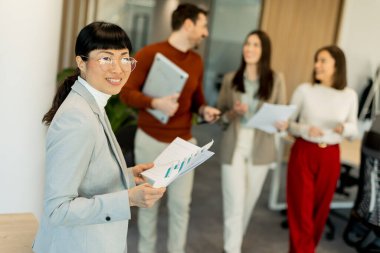 Gösterişli bir ofiste önemli belgeleri inceleyen odaklanmış bir profesyonel, meslektaşları arka planda tartışırken, işbirliği ve dinamik bir çalışma ortamı sunuyor..