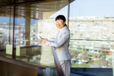 Parlak güneş ışığı şık takım elbiseli kendine güvenen Asyalı bir kadını aydınlatıyor, akıllı telefonuyla hareketli bir şehir ortamında çağdaş mimarinin arka planına karşı tebessüm ederken gülümsüyor..