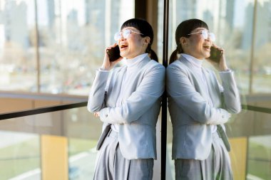 Modern ofis ortamında, şık takım elbiseli kendine güvenen Asyalı bir kadın telefonda konuşurken geniş bir gülümseme sergiliyor, yansıması şehrin cam zemininde görülebiliyor..