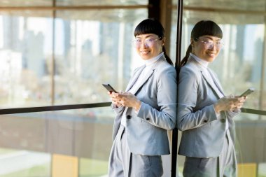 Şık bir iş elbisesi giymiş odaklanmış profesyonel bir Japon kadın akıllı telefonundan kayıyor. O, dışarıda canlı bir şehir manzarasını yansıtan büyük bir cam pencerenin yanında duruyor..