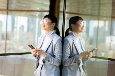 Şık bir iş elbisesi giymiş odaklanmış profesyonel bir Japon kadın akıllı telefonundan kayıyor. O, dışarıda canlı bir şehir manzarasını yansıtan büyük bir cam pencerenin yanında duruyor..