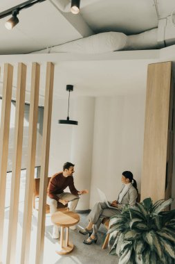 İki profesyonel, doğal ışıkla dolu modern bir çalışma alanında canlı bir tartışmaya girişiyor. Biri ahşap bir sandalyede otururken, diğeri dikkatle bir dizüstü bilgisayarla çalışıyor..