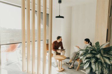 Çağdaş giyimli iki birey, ahşap aksanlar ve yeşillik içeren parlak bir salonda dikkatle sohbet ediyorlar. Güneş ışığı yumuşak gölgeler bırakır, tartışmalarına sıcaklık katar..