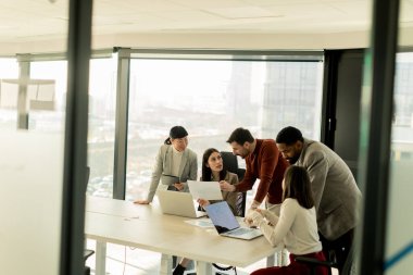 Çeşitli profesyonellerden oluşan bir grup, bir konferans masası etrafında dinamik bir beyin fırtınası yapıyor, fikirlerini paylaşıyor ve parlak bir ofis alanında dizüstü bilgisayar ve notlarla etkileşime giriyor..
