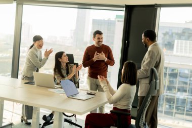 Bir grup profesyonel, geniş pencereleri ve doğal ışığı olan çağdaş bir ofiste proje planlarını aktif olarak tartışıyor. Müdahaleleri takım çalışması ve yenilik sürecini yansıtıyor.