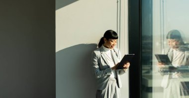 Gösterişli bir ofis alanında, profesyonel kıyafetli Japon kadın büyük bir pencerenin yanında duruyor, tabletiyle etkileşime giriyor, güneş ışığının sıcacık ışığında güneşleniyor, odaklanmış tavrını yansıtıyor..