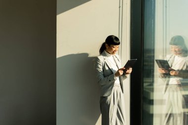 Modern bir ofiste büyük bir pencerenin yanında duran Japon iş kadını tabletini kullanıyor, işine odaklanıyor. Güneş ışığı aşağıdaki hareketli şehrin neşeli atmosferini aydınlatır..