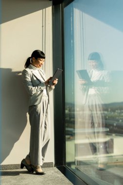 Modern bir ofiste büyük bir pencerenin yanında duran Japon iş kadını tabletini kullanıyor, işine odaklanıyor. Güneş ışığı aşağıdaki hareketli şehrin neşeli atmosferini aydınlatır..