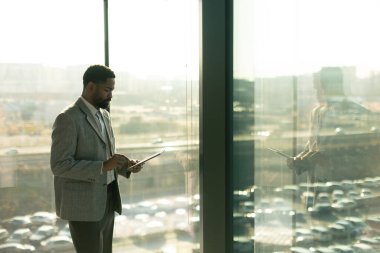 İyi giyimli bir adam parlak bir ofis alanında tablet kullanırken gülümser. Güneş ışığı sıcak bir parıltı saçar ve üretken bir anın tadını çıkarırken odaklanmış yüz ifadesini vurgular..