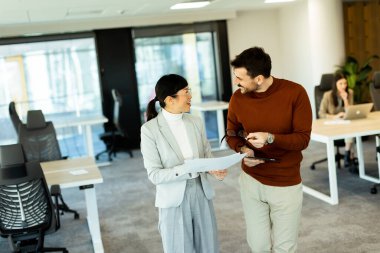İki profesyonel, çağdaş bir ofiste yürürken doküman ve fikir alışverişinde bulunuyorlar..