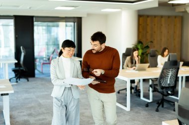 Işıl ışıl bir ofiste iki profesyonel iş birliği içinde hareketli bir değişim gerçekleşiyor. Arka planda, iş arkadaşları dizüstü bilgisayarlarına odaklanarak takım çalışması ve yenilik atmosferini artırıyorlar..