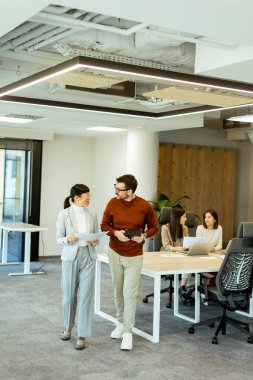 Işıl ışıl bir ofiste iki profesyonel iş birliği içinde hareketli bir değişim gerçekleşiyor. Arka planda, iş arkadaşları dizüstü bilgisayarlarına odaklanarak takım çalışması ve yenilik atmosferini artırıyorlar..