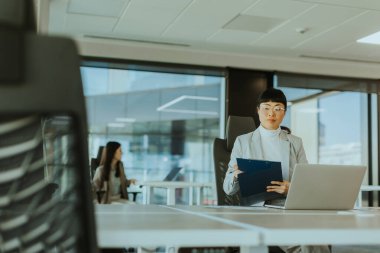 Gösterişli bir ofiste, odaklanmış Asyalı bir kadın masasındaki belgeleri incelerken, bir meslektaşı da yakınlarda iş birliği ile dolu dinamik bir çalışma ortamı sergiliyor..