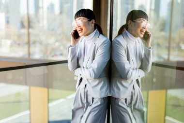 Modern ofis ortamında, şık takım elbiseli kendine güvenen Asyalı bir kadın telefonda konuşurken geniş bir gülümseme sergiliyor, yansıması şehrin cam zemininde görülebiliyor..