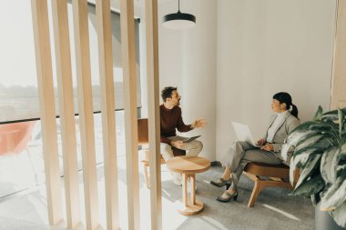 İki profesyonel, doğal ışıkla dolu modern bir çalışma alanında canlı bir tartışmaya girişiyor. Biri ahşap bir sandalyede otururken, diğeri dikkatle bir dizüstü bilgisayarla çalışıyor..