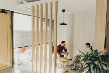 Çağdaş giyimli iki birey, ahşap aksanlar ve yeşillik içeren parlak bir salonda dikkatle sohbet ediyorlar. Güneş ışığı yumuşak gölgeler bırakır, tartışmalarına sıcaklık katar..