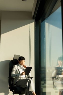 Güneşli bir ofis alanında, resmi giyinmiş Japon iş kadını modern bir sandalyede rahatça oturur. Dikkatle tabletine odaklanıyor, hırsını ve konsantrasyonunu yansıtıyor..