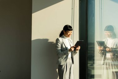 Gösterişli bir ofis alanında, profesyonel kıyafetli Japon kadın büyük bir pencerenin yanında duruyor, tabletiyle etkileşime giriyor, güneş ışığının sıcacık ışığında güneşleniyor, odaklanmış tavrını yansıtıyor..