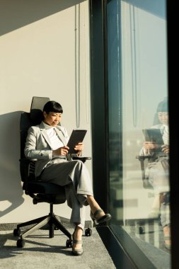 Güneşli bir ofis alanında, resmi giyinmiş Japon iş kadını modern bir sandalyede rahatça oturur. Dikkatle tabletine odaklanıyor, hırsını ve konsantrasyonunu yansıtıyor..