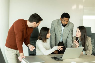 Parlak bir ofiste, çeşitli profesyonellerden oluşan bir grup dizüstü bilgisayarın etrafında toplanarak, dinamik tartışmalar, takım çalışması ve yaratıcılıkla uğraşırken içgörü ve stratejilerini paylaşıyor..