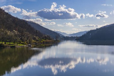 Parlak mavi bir gökyüzünün altında, gür yeşillik suyu sararken huzurlu bir göl kabarık bulutları yansıtır. Yumuşak tepeler ufku sarar, doğada sakin bir kaçış yaratır..