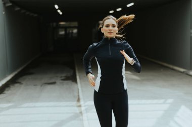 Şık bir spor kıyafetle giyinmiş bir kadın şık bir şehir tüneline doğru koşuyor. Sabah ışığında enerji ve kararlılığı somutlaştırıyor..