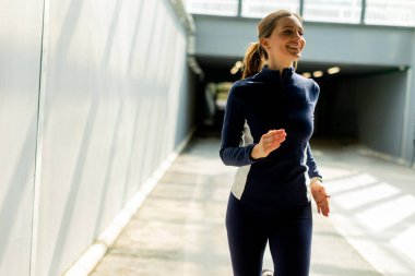 Atletizm kıyafetleri giymiş bir kadın şık bir şehir yolunda güvenli bir şekilde koşuyor. Güneş ışığı binanın tasarımını süzerken gülümsüyor..