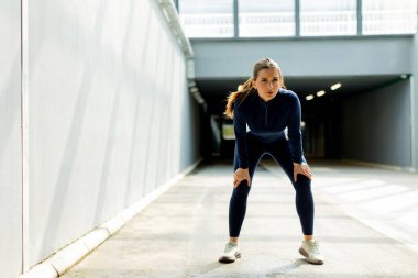 Odaklanmış ve kararlı bir şekilde, atletik giyinen bir kadın ellerini dizlerinin üstüne çöküp, kendini zorladıktan sonra pırıl pırıl, güneşli bir koridorda iyileşiyor..
