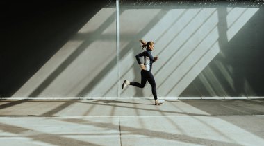 Bir kadın, aydınlık bir sabahta yakındaki binalardan gelen gölgelerle aydınlanan beton bir yolda enerjik bir şekilde koşar..
