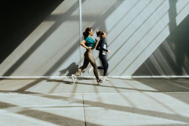 İki kadın kararlı bir şekilde modern bir yapıdan yayılan yumuşak sabah ışığıyla aydınlatılarak çarpıcı gölgeler oluşturarak ileriye doğru koşar..