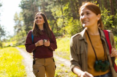 İki arkadaş yemyeşil bir ormanda serinletici bir yürüyüşün tadını çıkarıyorlar, ellerinde kupalar ve doğanın güzelliklerindeki neşeli anları yakalıyorlar..