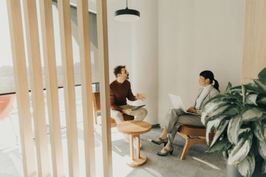 İki profesyonel, doğal ışıkla dolu modern bir çalışma alanında canlı bir tartışmaya girişiyor. Biri ahşap bir sandalyede otururken, diğeri dikkatle bir dizüstü bilgisayarla çalışıyor..