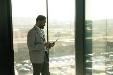 İyi giyimli bir adam parlak bir ofis alanında tablet kullanırken gülümser. Güneş ışığı sıcak bir parıltı saçar ve üretken bir anın tadını çıkarırken odaklanmış yüz ifadesini vurgular..